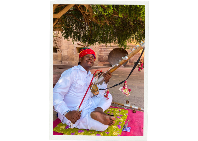 Smiling musician in traditional attire playing a Rajasthani instrument outdoors.
