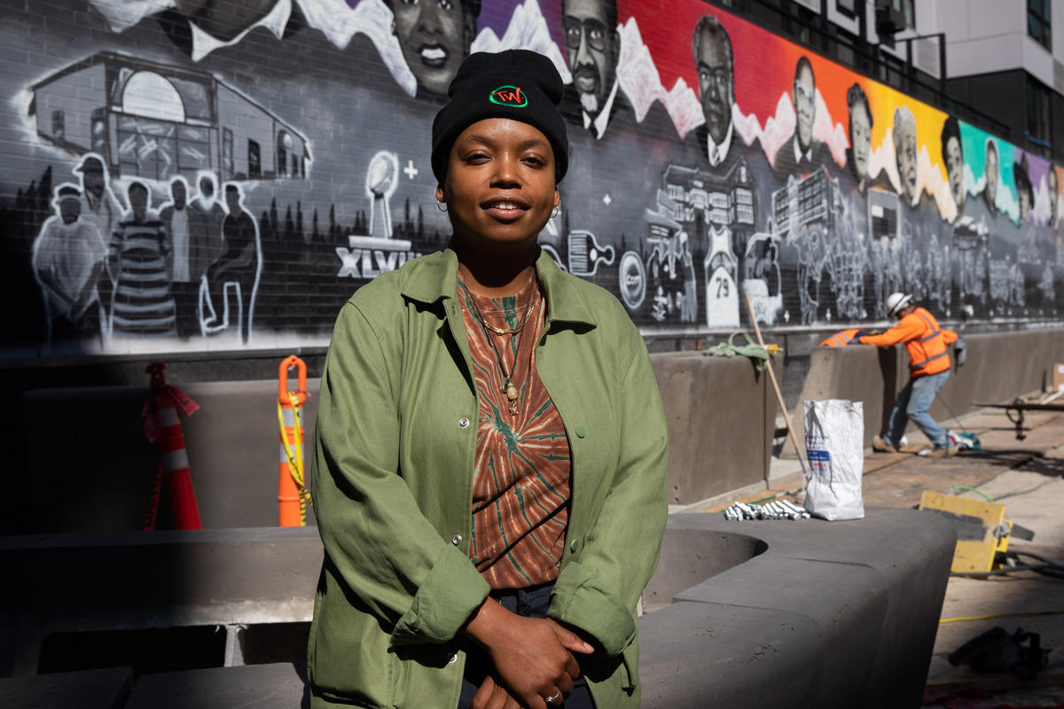 A black woman poses in front of a street mural depicting Black historical figures, buildings, and other cultural items.