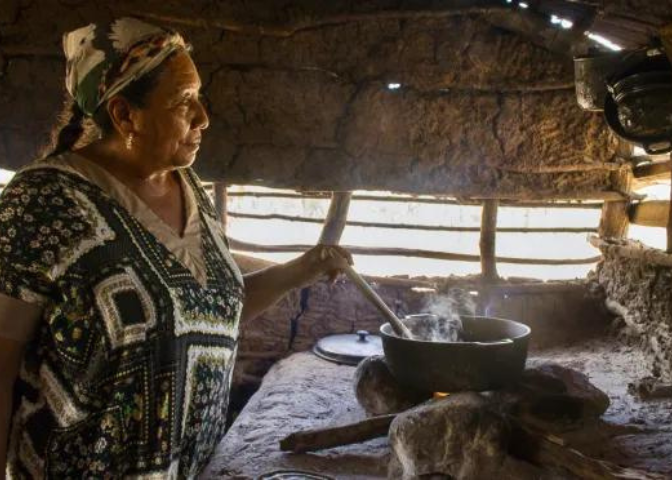 Photo depicting an Indigenous elder woman preparing something in a pot over a wood-fire stove.