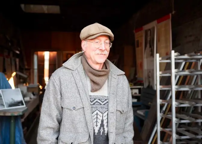 A man stands in a dark hallway. He is wearing a hat, scarf, and coat.