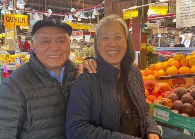 A husband and wife smile and stand in front of a fruit stand at Pike Place Market.
