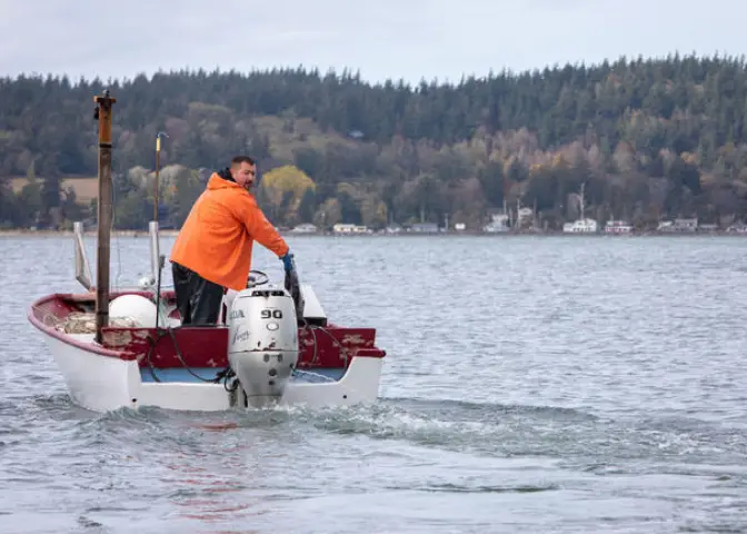 A fisherman in an orange raincoat stands in a boat, speeding across the water.