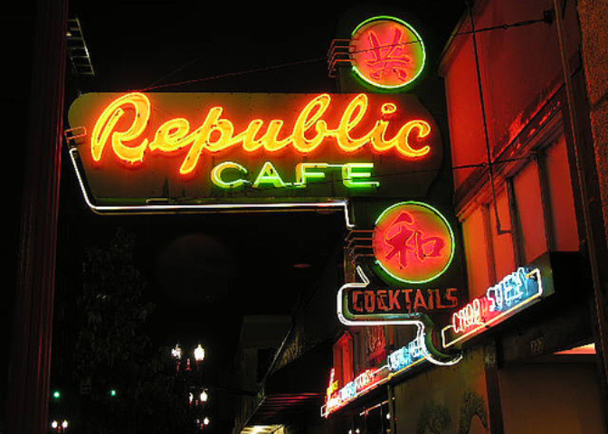 A neon sign of the Republic Cafe is illuminated on a busy street at night.