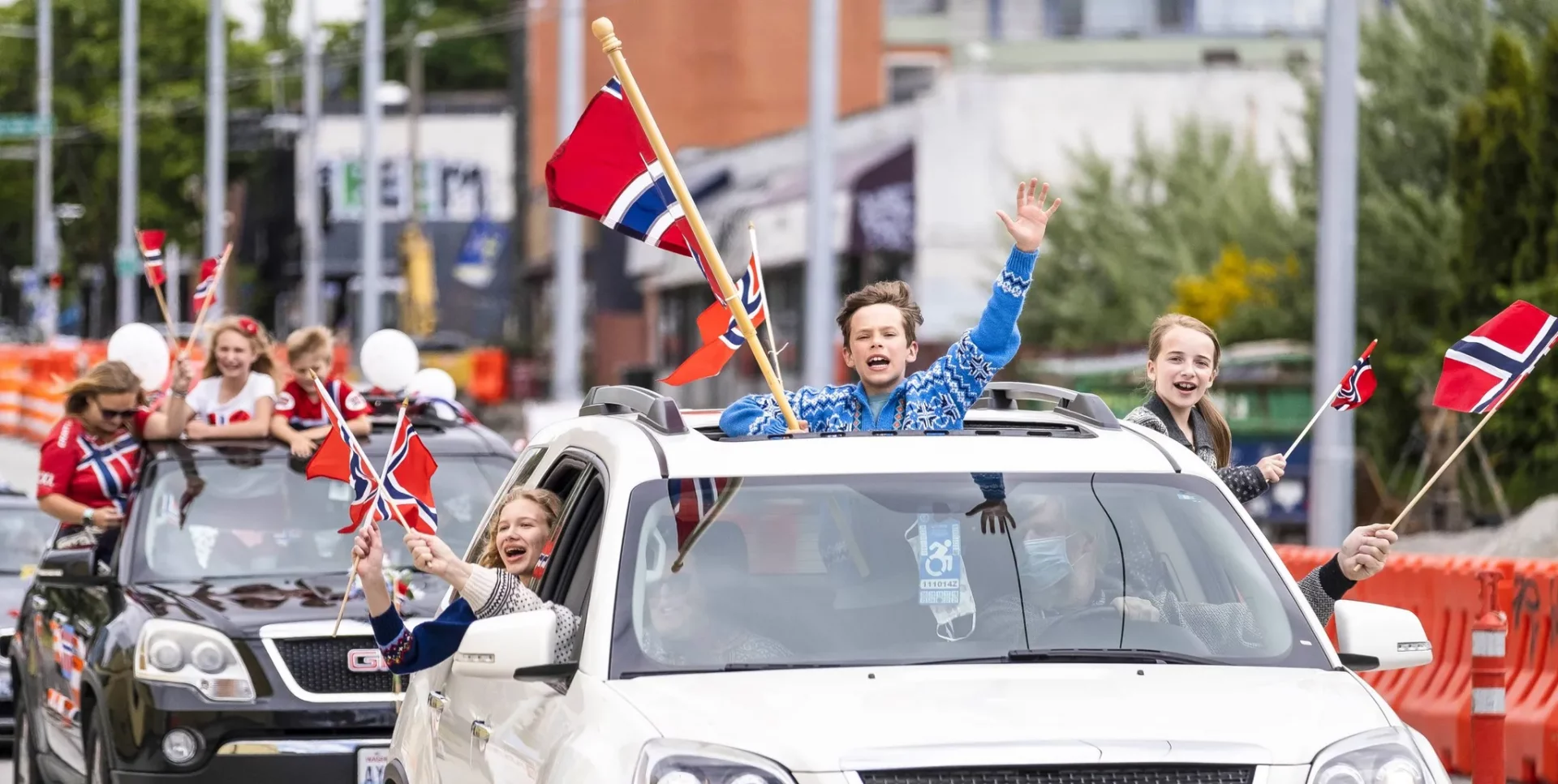 A boy sticks his head out the sunroof of a car in a parade, waving and holding a Norwegian flag.