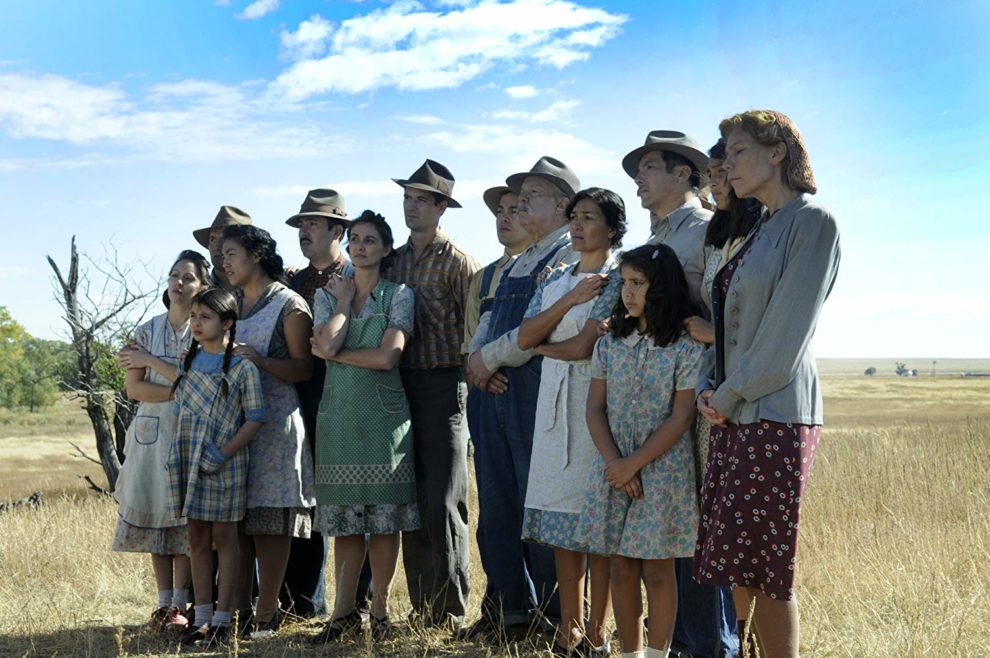 A group posing for a photo in a brown grass field. The women wear dresses, the men wear buttoned shirts and Stetson hats.