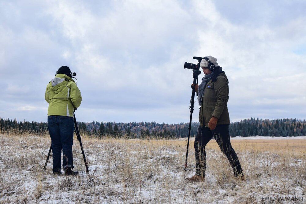 Two people in heavy coats position cameras on a large field dusted with snow.