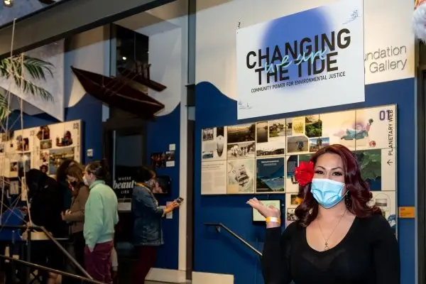 A woman stands in front of an exhibit at a gallery titled "We Are Changing the Tide".