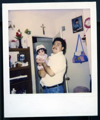 A moustached father stands in the middle of a kitchen holding his infant daughter.