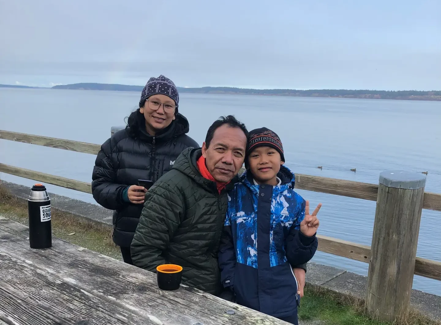 A mother, father, and son wearing winter coats drink tea from a thermos at a picnic table by the beach.