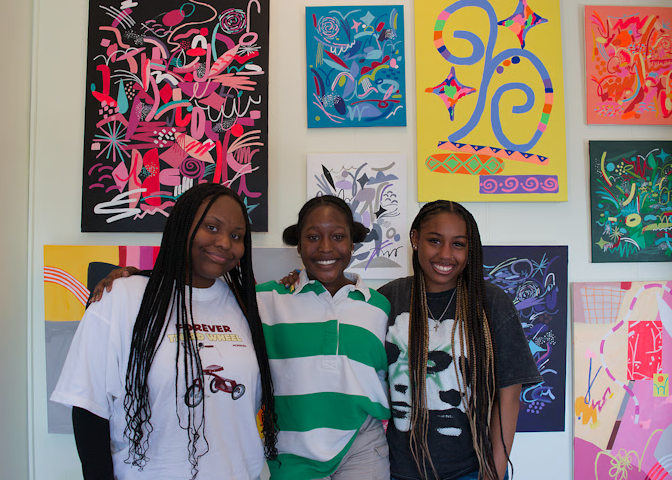 Three young Black women stand shoulder to shoulder in front of a wall of colorful paintings.