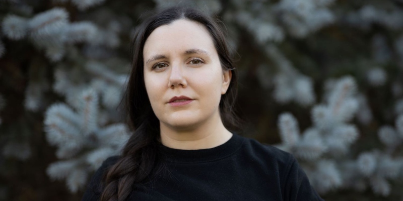 A woman, dressed in black, looks at the camera in front of some pine trees.