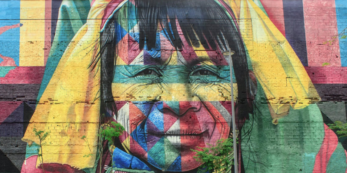 A mural painted on a brick wall of an Indigenous woman in a wide spectrum of colors.
