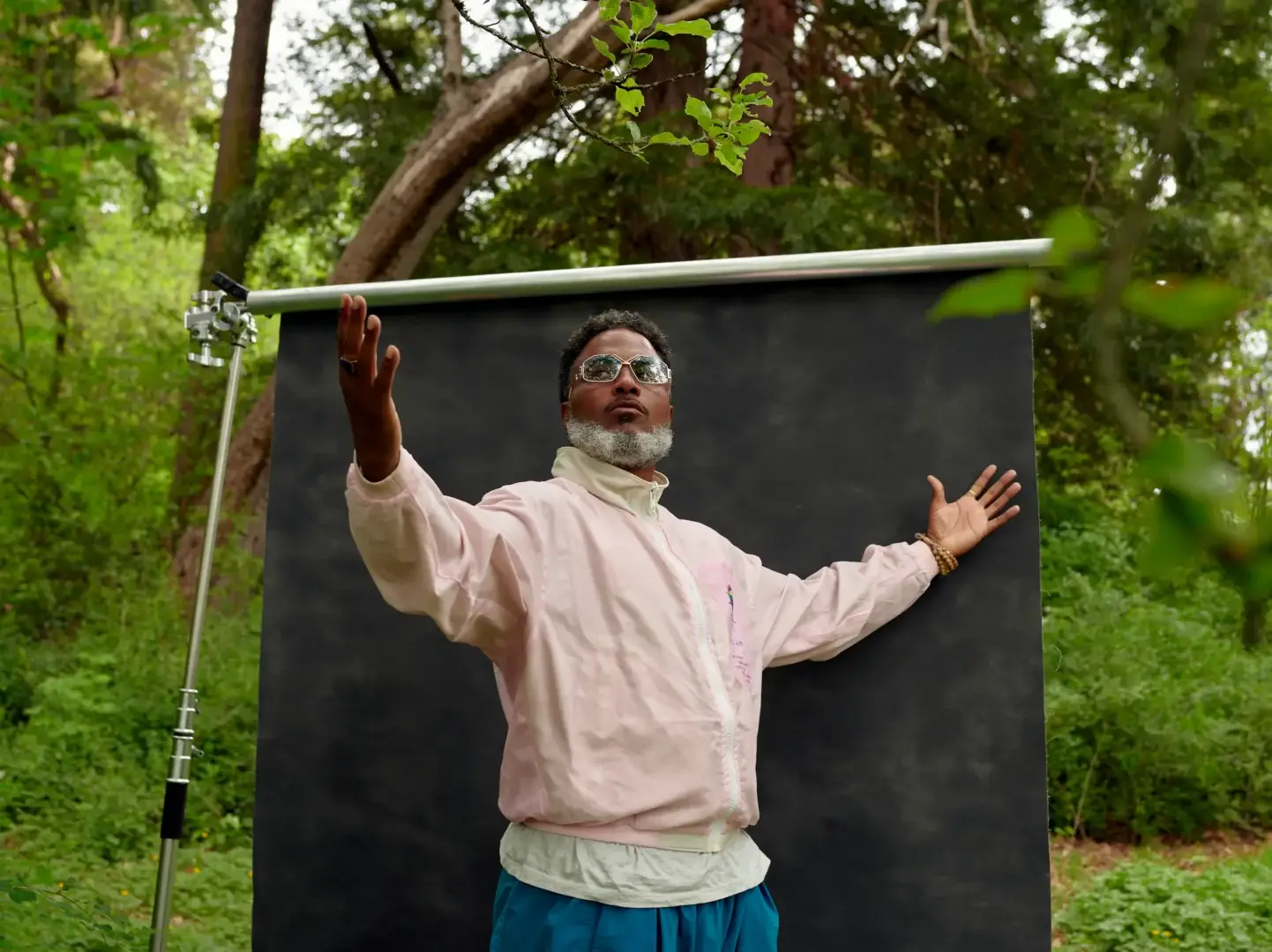 A man in a turtleneck with arms outstretched poses for a portrait surrounded by trees.