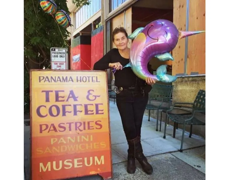A woman in black stands with a narwhal balloon on a sidewalk next to a sign that says 'Panama Hotel Tea & Coffee'