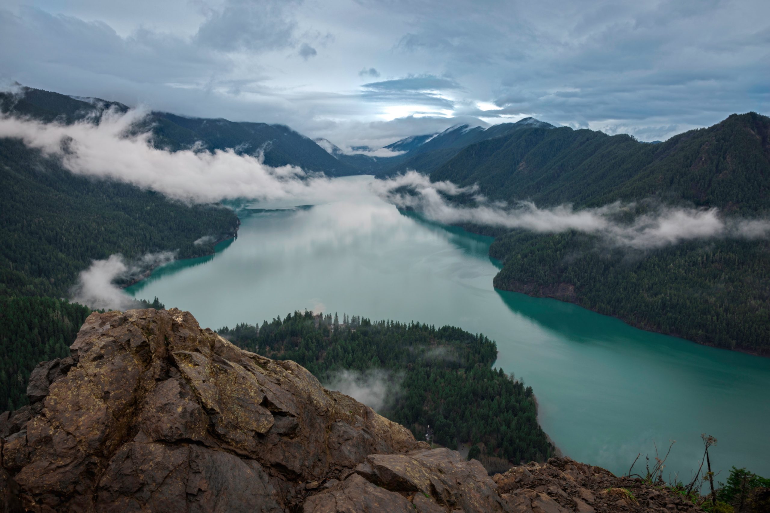 A view from atop a hill overlooking a vast and cloudy lake surrounded by forest and hills.