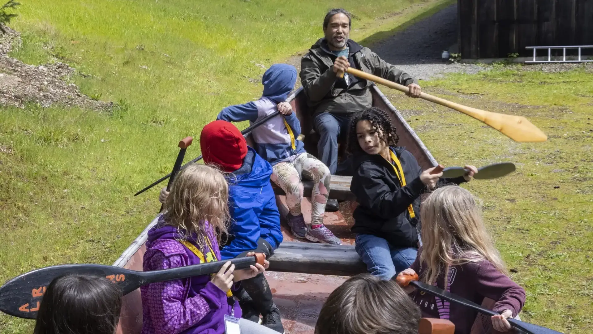 A man teaches a group of children to row in a canoe on the grass.