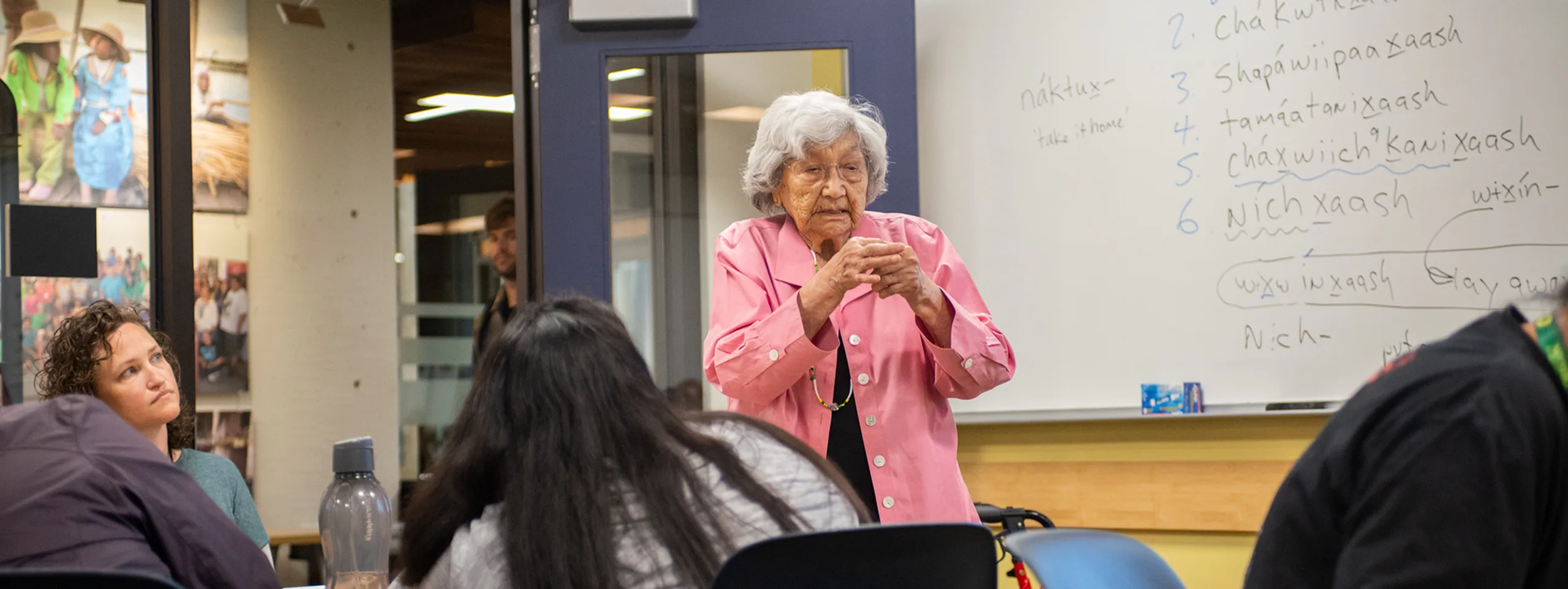 An elderly woman speaks in front of a classroom. On the whiteboard are words in an Indigenous language.
