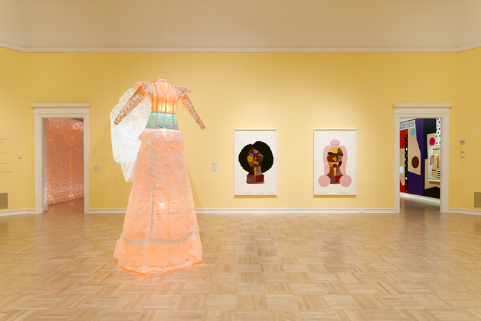 A large, pink dress stands in a room beside two portraits of Black women.