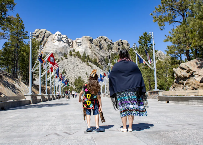 A woman and a young boy in traditional Native American clothing looking up at Mt. Rushmore in the background.