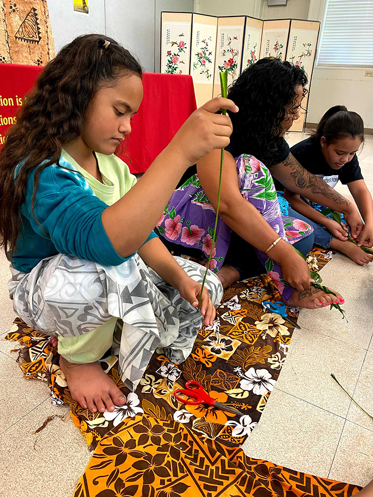 Young girls sitting on the floor threading leaves onto leis.