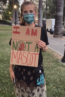 Koli Kohler wears a ribbon skirt and mask and holds an I Am Not A Mascot sign.
