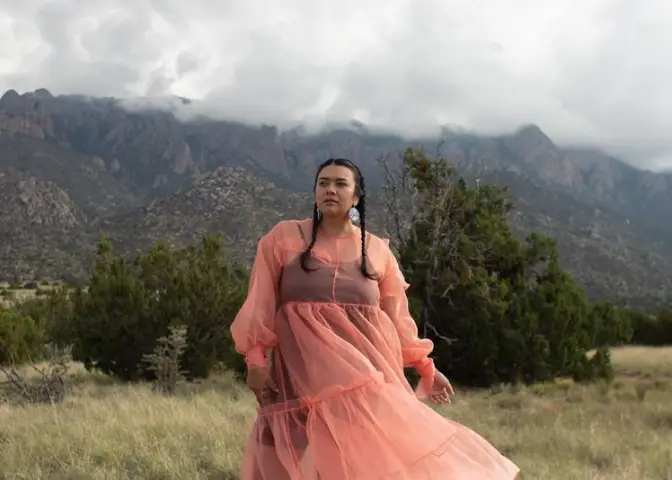 A woman with braids stands in a field with trees and bushes behind her.