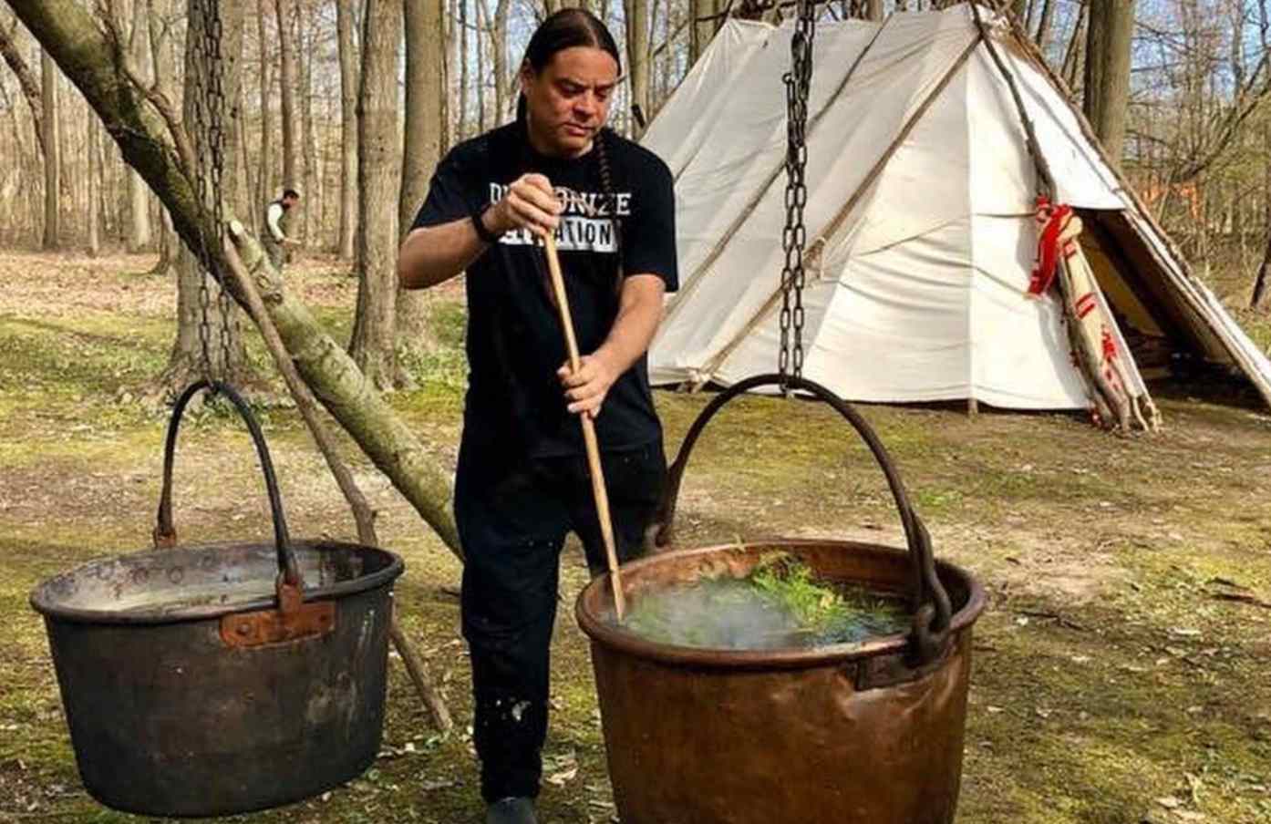 In a forest, a large pot is suspended by a chain over a wood fire while a man stirs it with a wooden spoon. In the background is a large traditional Native tent.