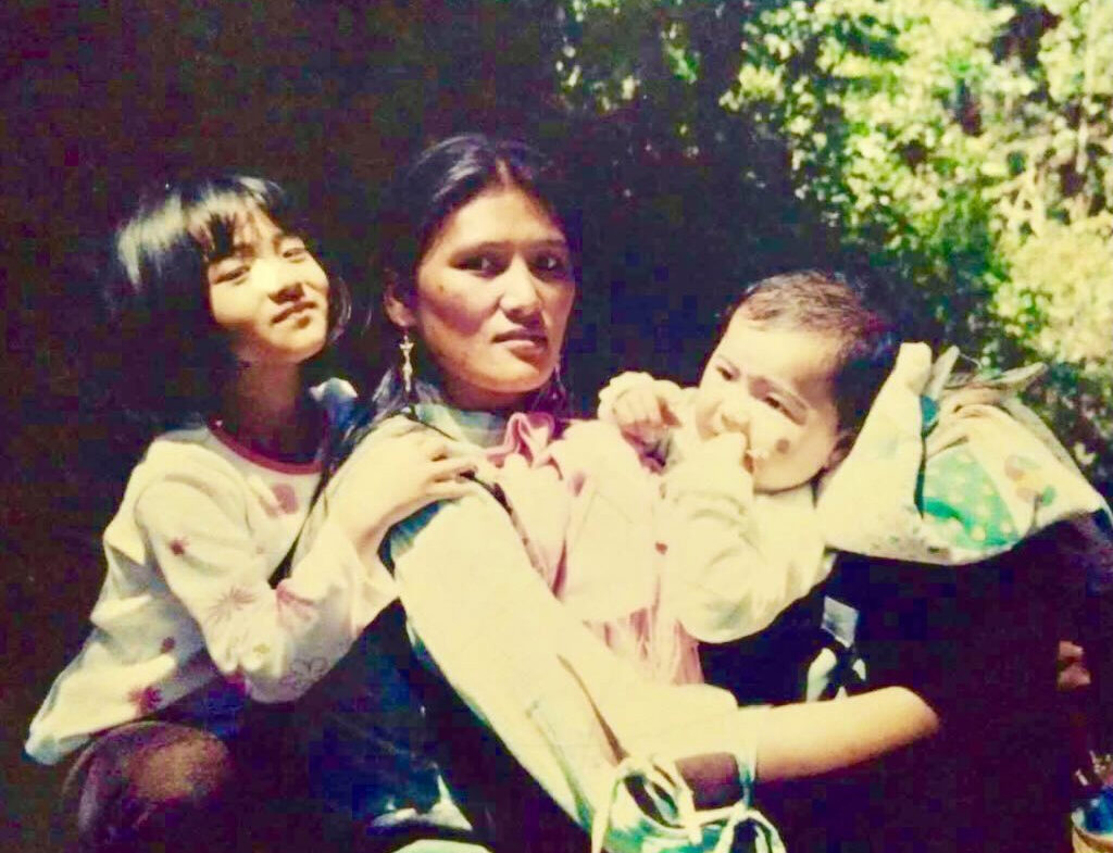 A mother holds her baby daughter in her arms, her older daughter hugs her from behind.