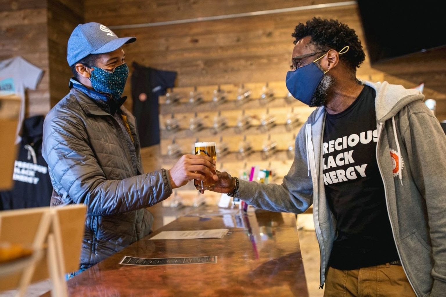 A black man hands a glass of beer to another black man in a brewery.