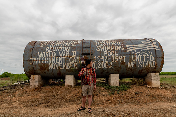 A man carrying a guitar over his shoulder in front of a tube decorated in remembrance of a World War II internment camp reunion.