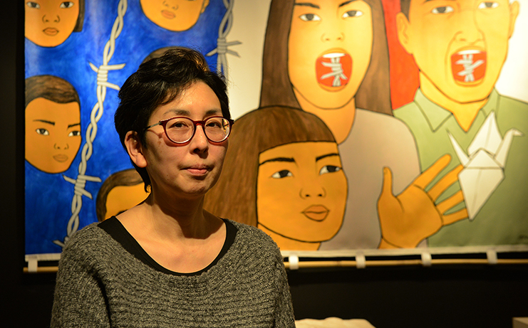 A woman in glasses poses in front of her painting of Japanese American pioneers before and during the incarceration of Japanese Americans in WW2.