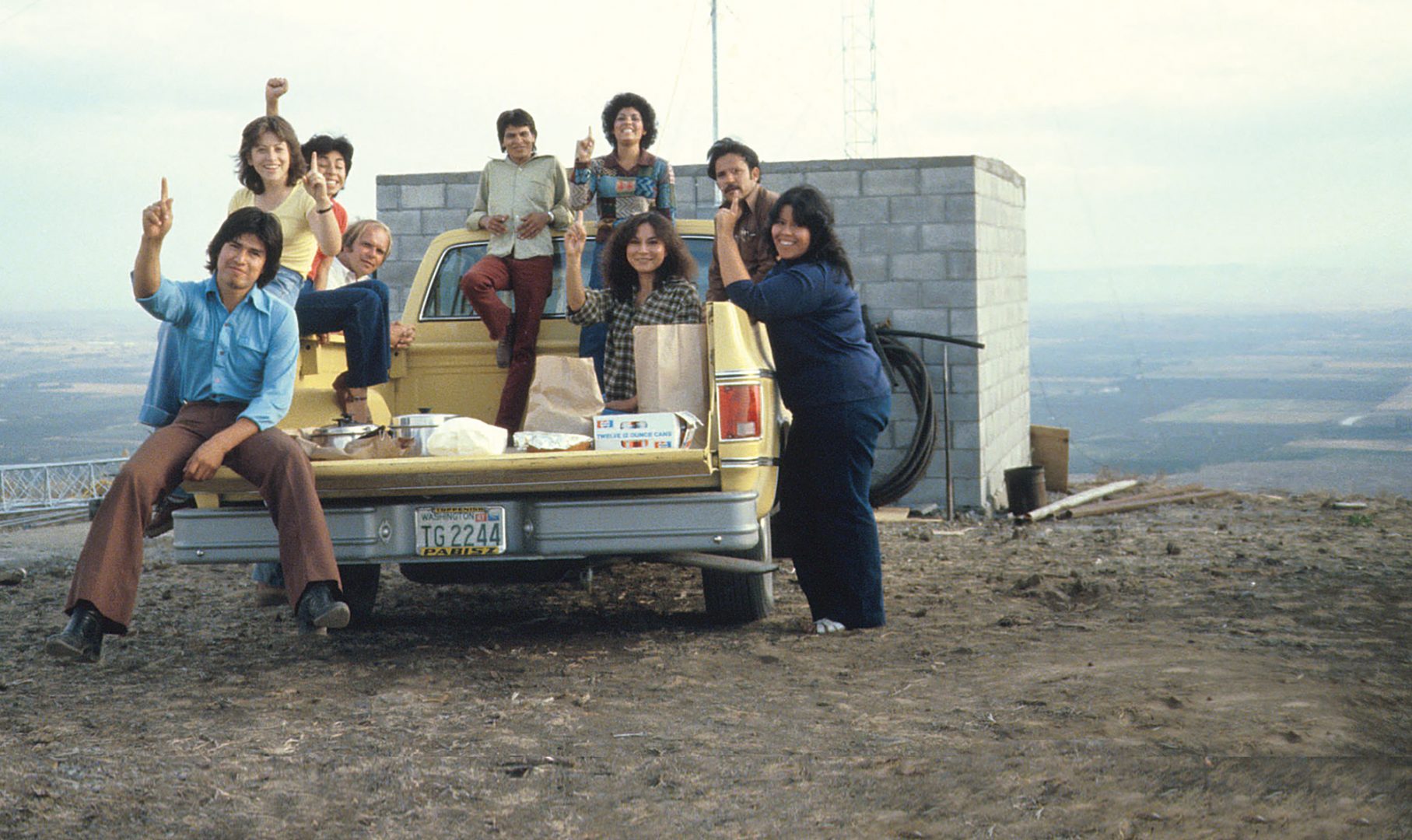 A group of people sitting on a truck in front of a small brick building overlooking a valley.