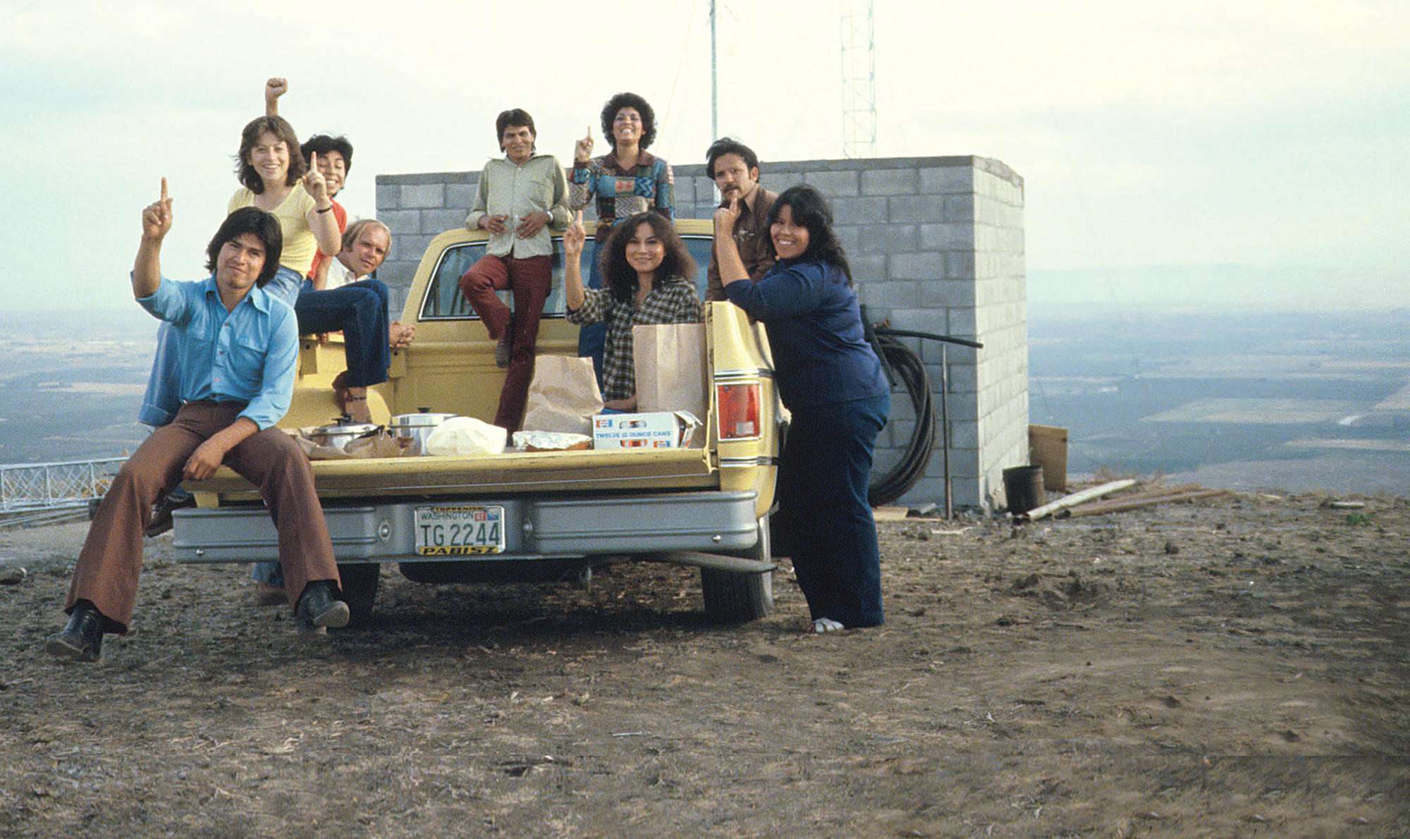 A group of people sitting on a truck in front of a small brick building overlooking a valley.